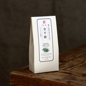 A white paper bag of Japanese Kompetto Sugar Candy standing on a rustic wooden surface against a dark brown background.